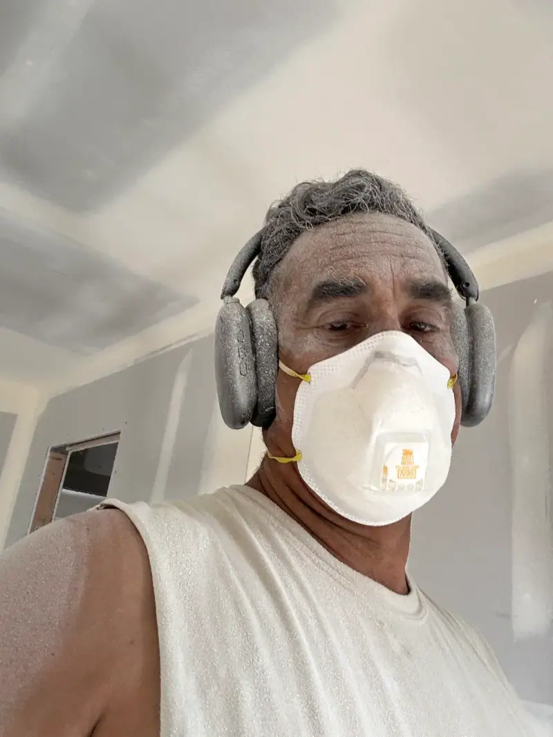 Danny of Danny Patches Custom Drywall in a respirator and ear protection, covered in sanding dust