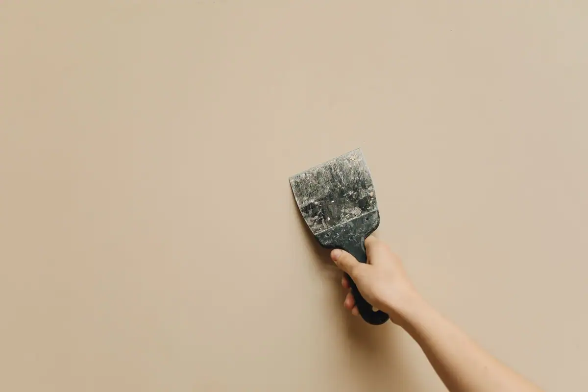 Hand holding a putty knife against a tan wall